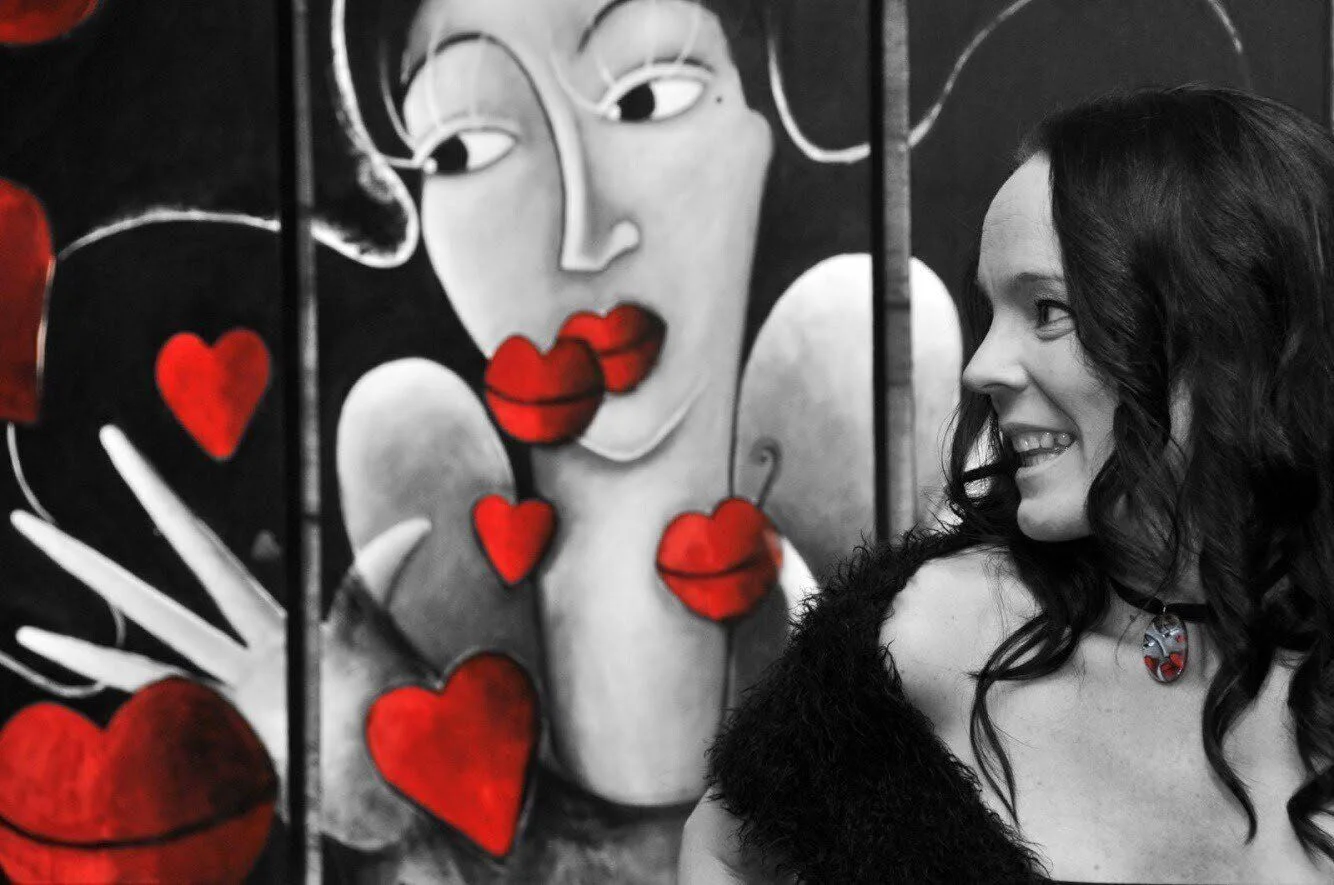 Nuria Catedra sonríe junto a uno de sus cuadros: una figura en blanco y negro rodeada de corazones y labios rojos.